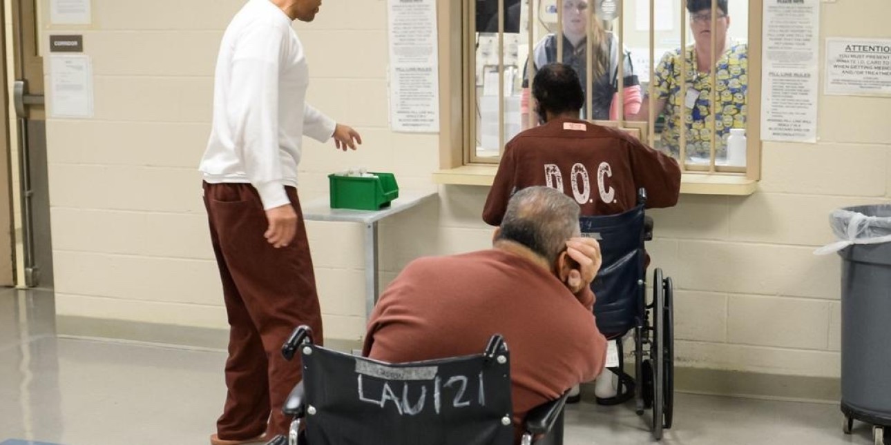 a photo shows two elderly incarcerated people in wheelchairs in line for medication and one incarceraeted person in a white shirt standing next to them