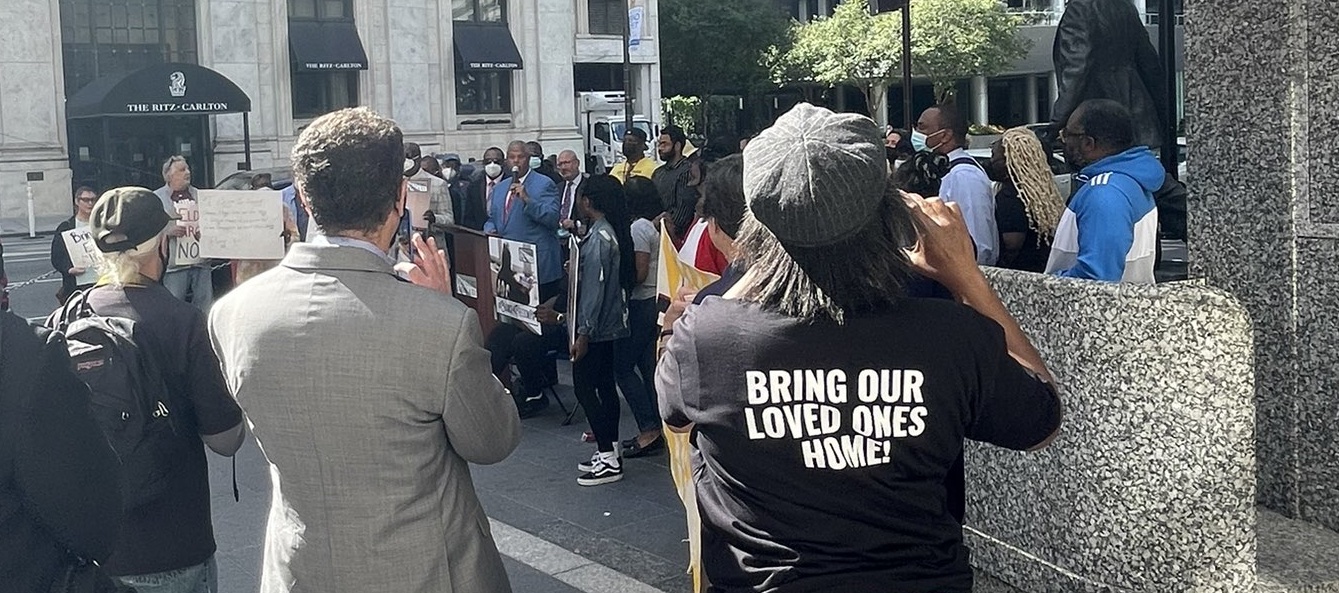 a woman wears a t-shirt that reads 'Bring Our Loved Ones Home' and looks on at a press conference