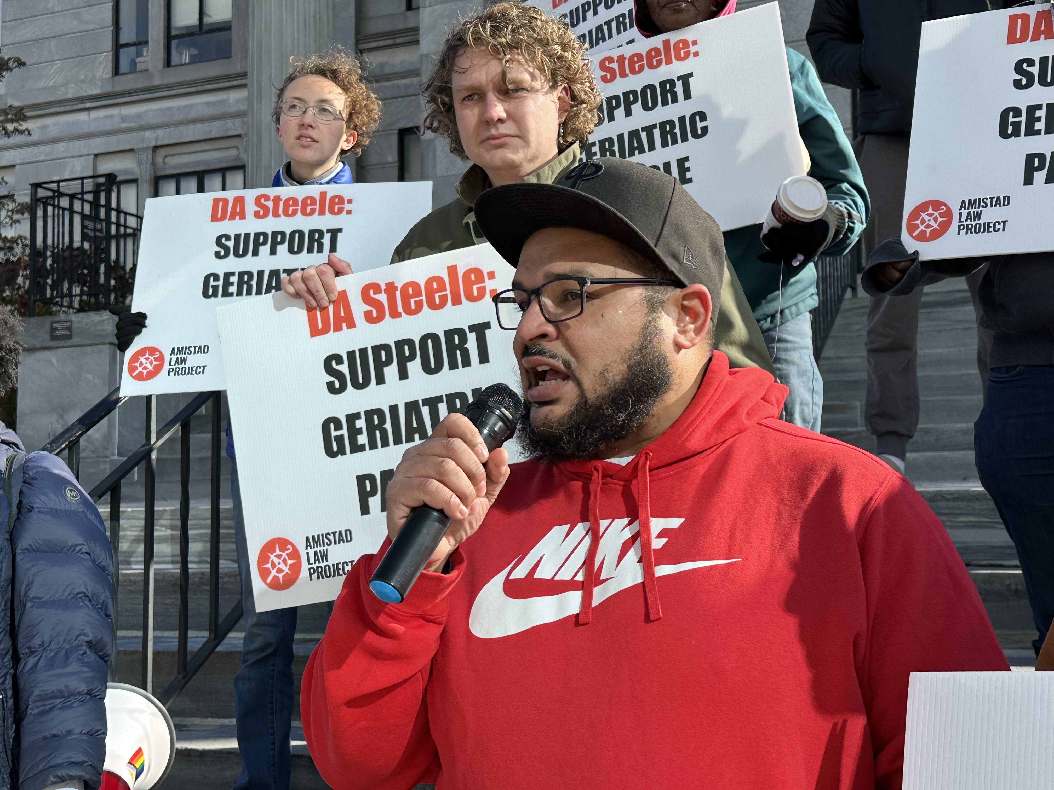 Sergio Hyland speaks into the mic surrounded by people with signs that read 'DA Steele Support Geriatric Parole.'