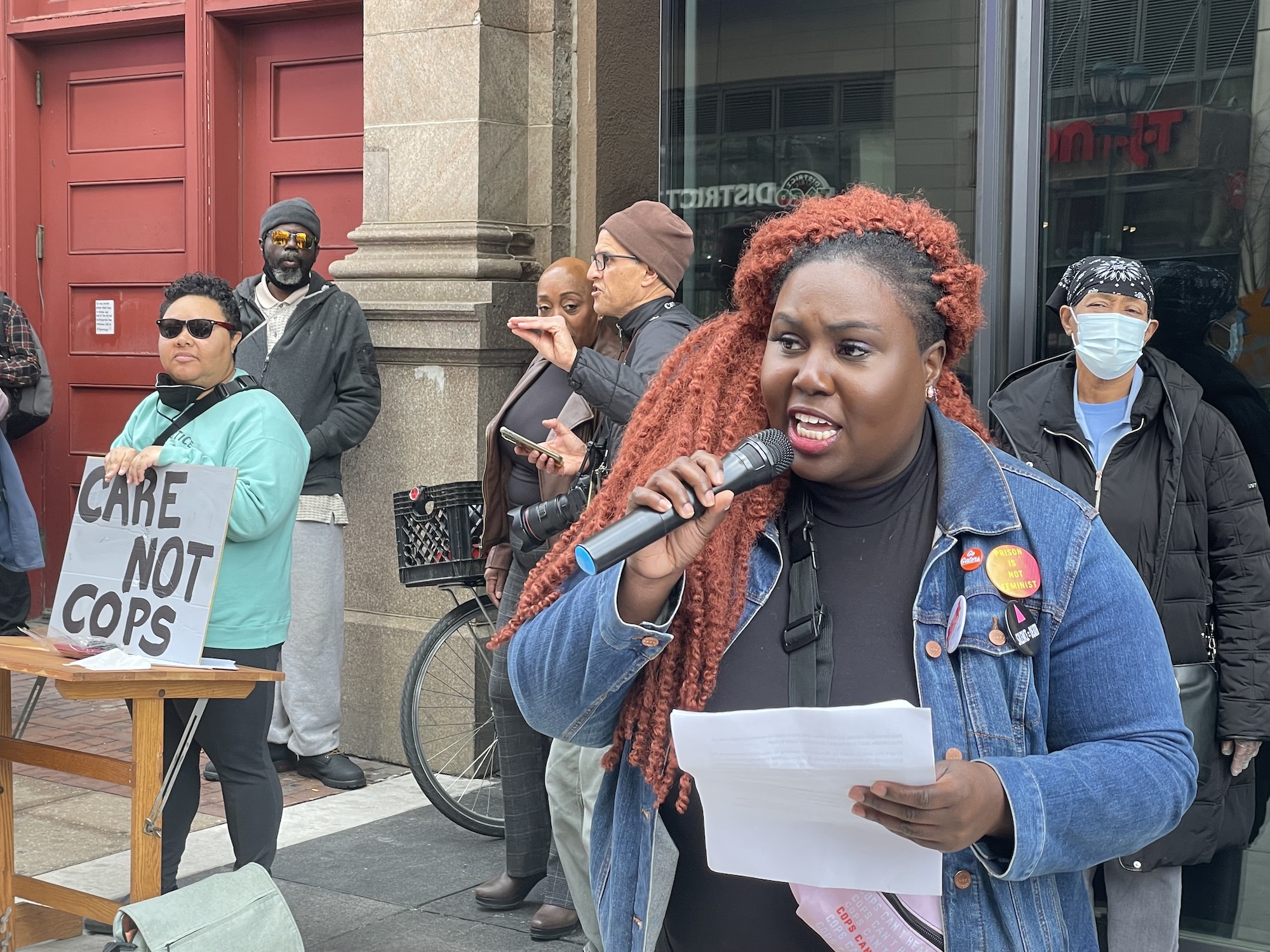 Nikki Grant holds a mic as Kris Henderson stands nearby with a sign that reads Care Not Cops. A crowd is gathered behind her