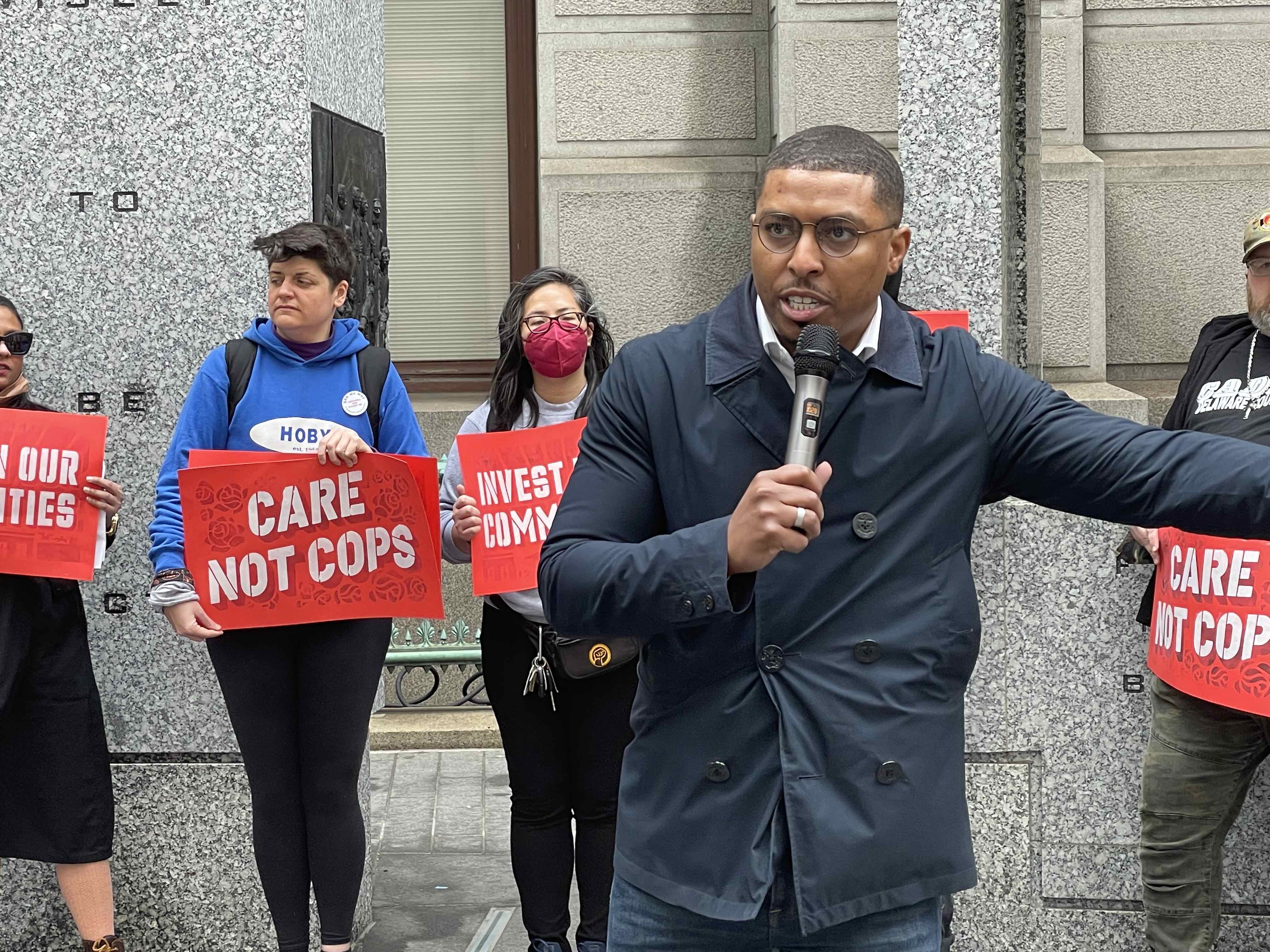 Nic O'rouke speaks to the crowd while people in the background hold sings that read 'Invest in Our Communities' and 'Care Not Cops'