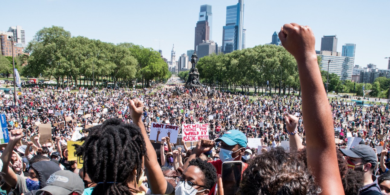 A rally of thousands of people assemble. The philly skyline is in the background and a Black person raises their fist in the foreground.