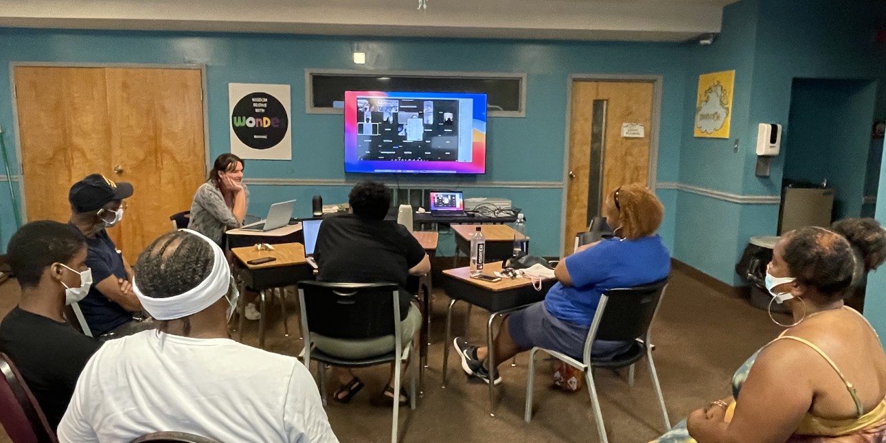 A group of family members of the incarcerated sit in chairs facing a screen where a bunch of people are online to join the in-real life participants in a hybrid meeting