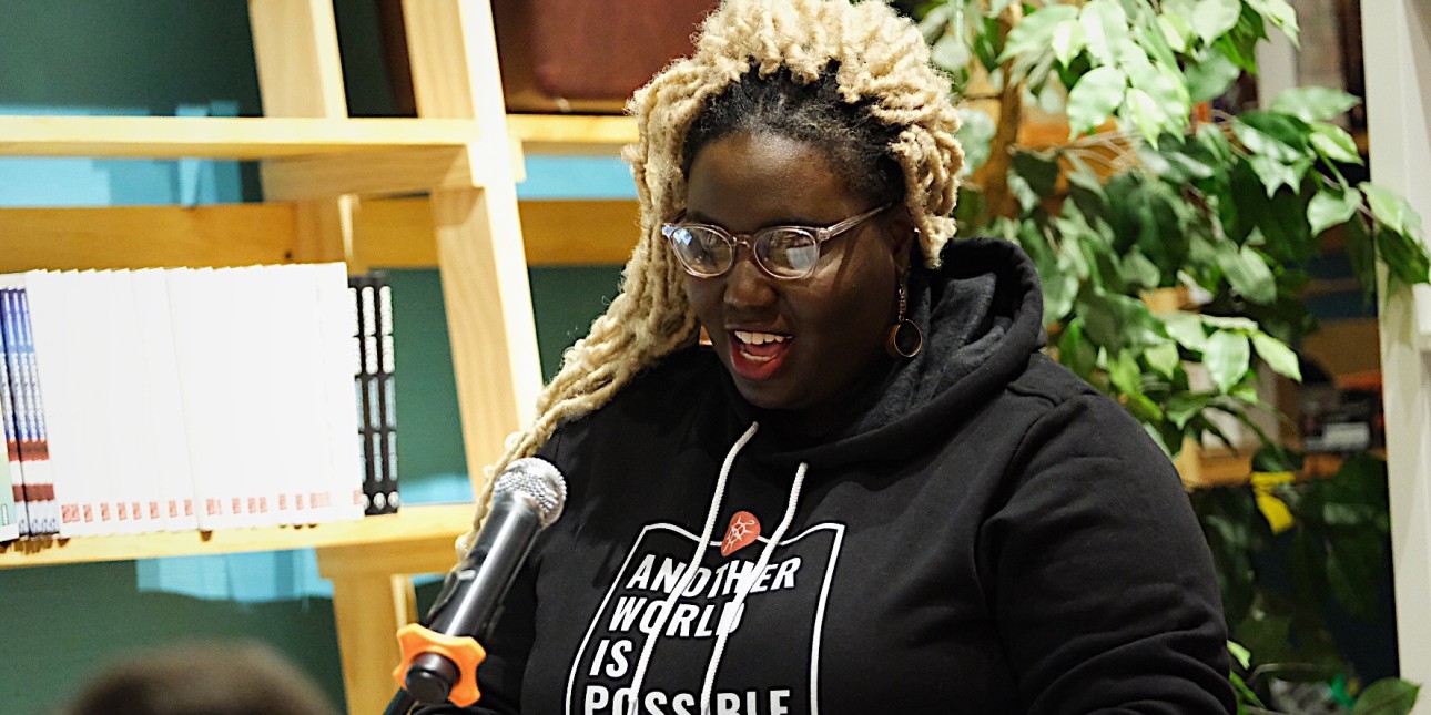 A Black woman with blond hair stands in front of a green plant and speaks into a microphone while wearing a black hooded sweatshirt with the words Another World is Possible on the front of it