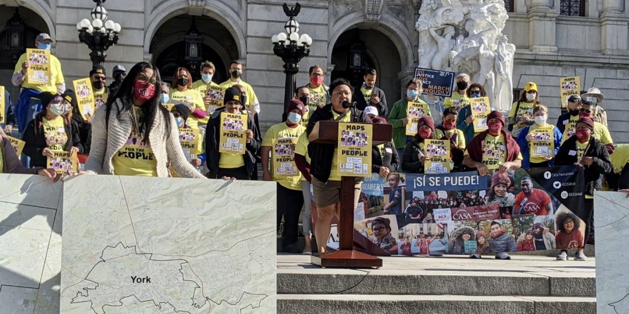 Amistad Law Project Executive Director Stands at a podium speaking  surrounded by people in yellow shirts in front of the state capitol building in Harrisburg