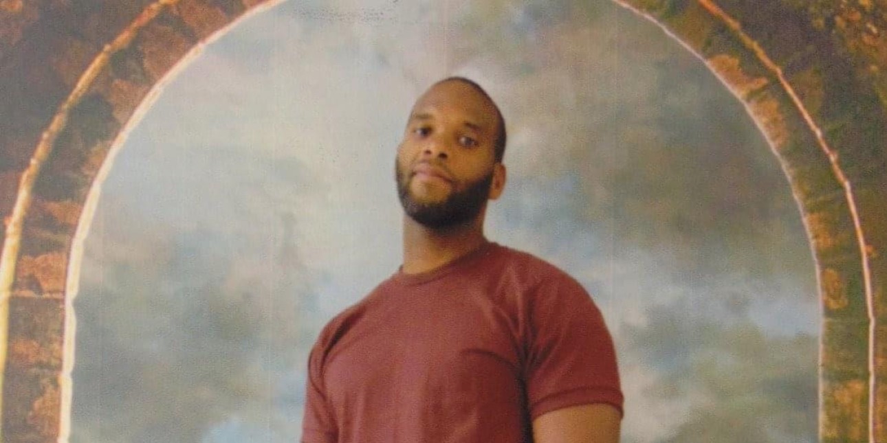 Clinton Walker wears a maroon colored shirt and looks down at the camera with an arch way behind him