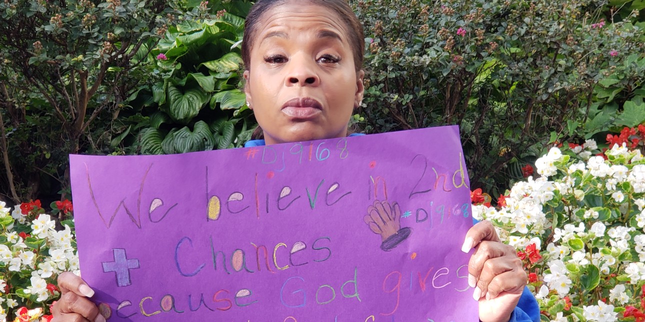 A Black woman looking with determination at the camera holds a purple sigh that says 'I believe in second chancess'