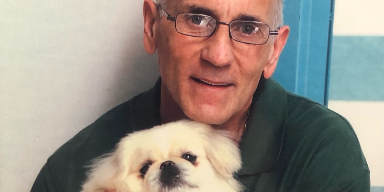 Richard Marra wears a green shirt and holds a small beige colored dog with a black nose and dark eyes