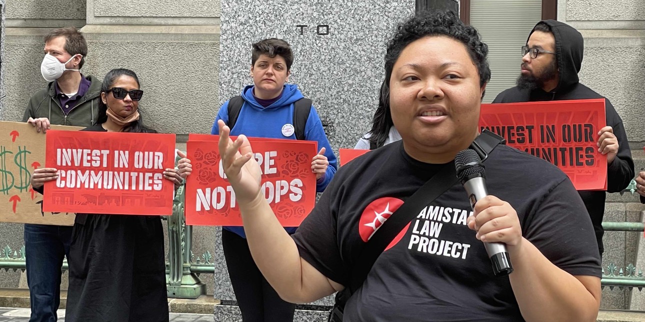 Kris Henderson holds a mic while people in the background hold signs that read 'Invest in Our Communities' and 'Care Not Cops'