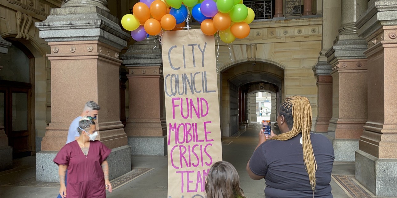 4 people stand around a banner held aloft by colorful balloons. The banner reads "City Council: Fund Mobile Crisis Teams NOW"