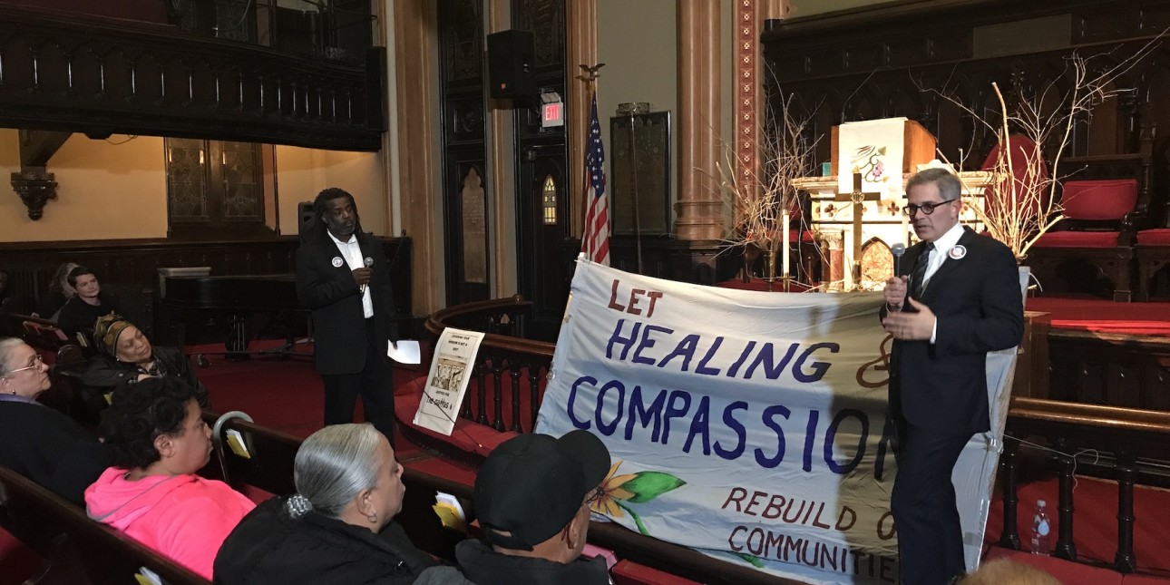 Larry Krasner stands in front of a banner that reads 'Healing and Compassion' in front of a mostly Black audience. 