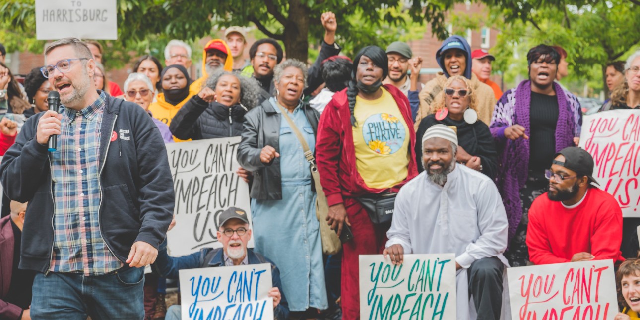 a crowd holds signs that read 'you can't impeach us' while sean damon of Amistad Law speaks on the mic