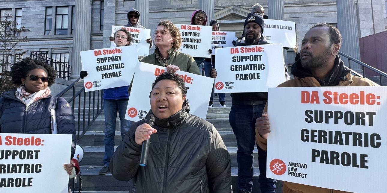 Kris Henderson speaks into a mic surrounded by people with signs that read 'DA Steele: Support Geriatric Parole'