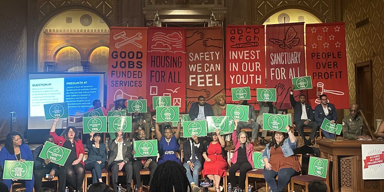 candidates for city council hold up green signs that say yes with red banners behind them 