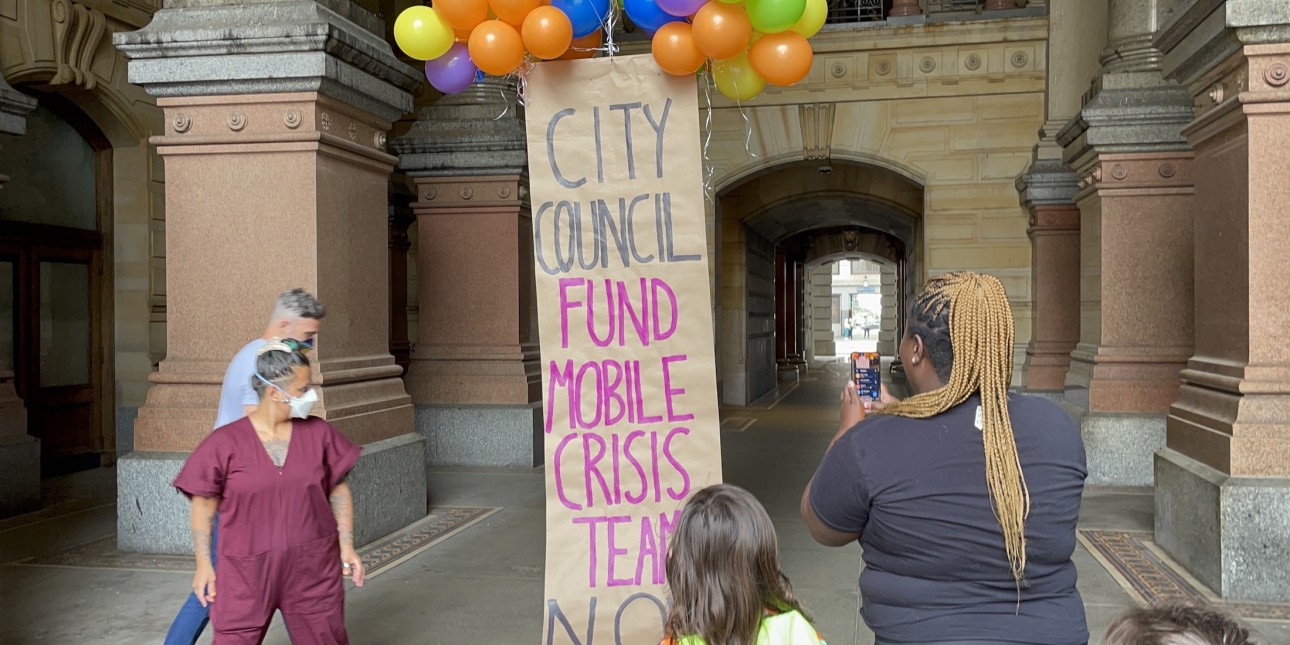 A banner lifted up by balloons reads 'City Council Fund Mobile Crisis Teams Now'