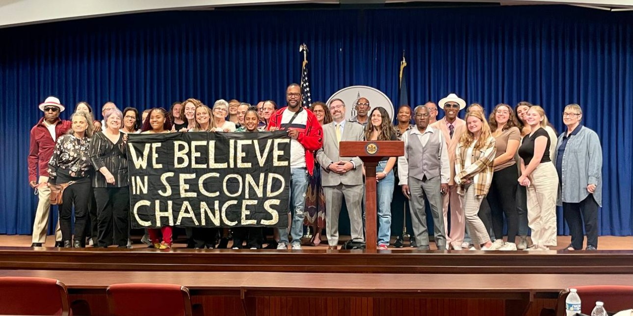 A couple dozen people stand on a stage with a banner that reads "we believe in second chances"