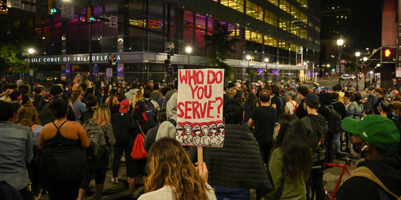 Crowd marches at protest with sign that reads "who do you serve?"