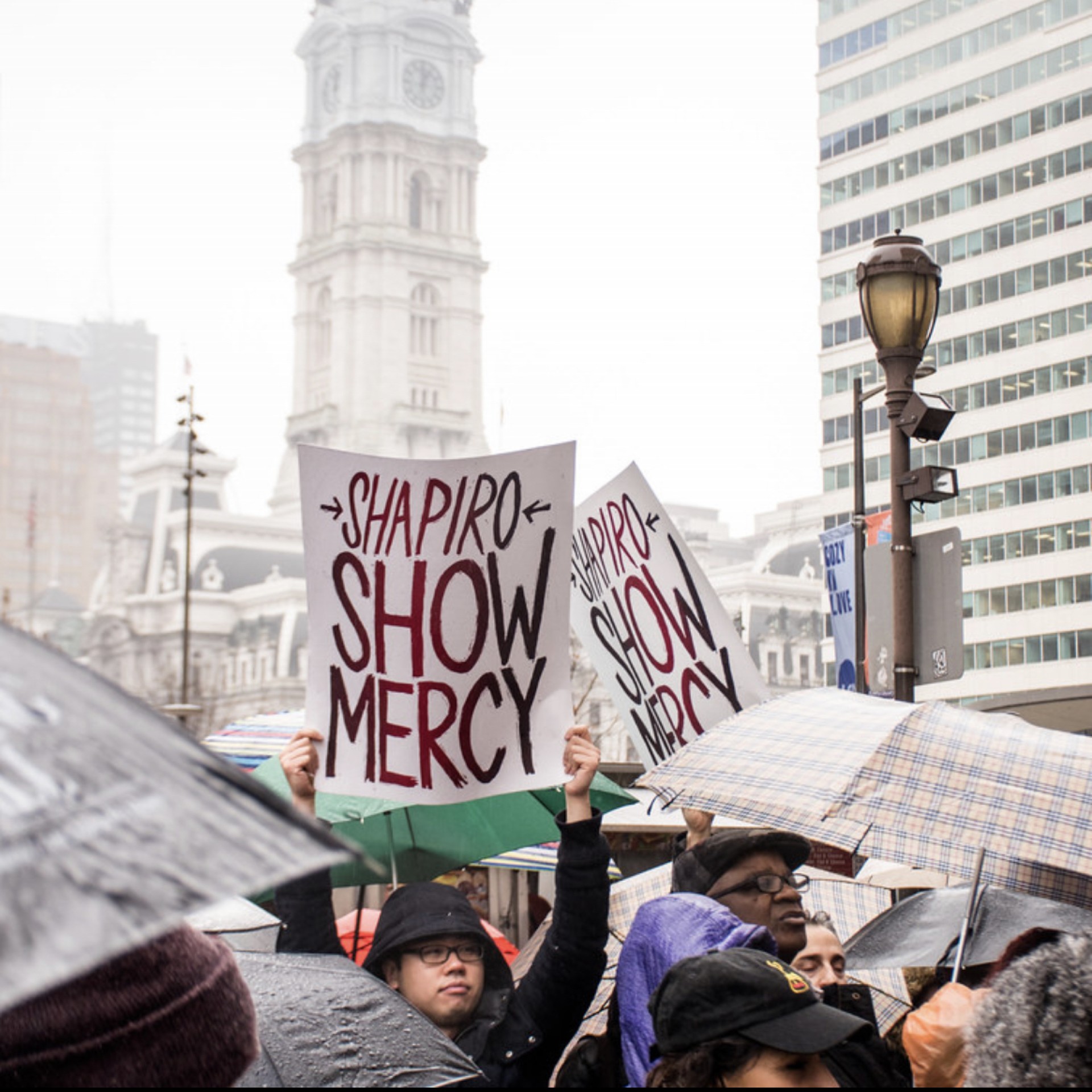 A person at a protest holds a sign that says 'Shapiro Show Mercy' in a sea of umbrellas