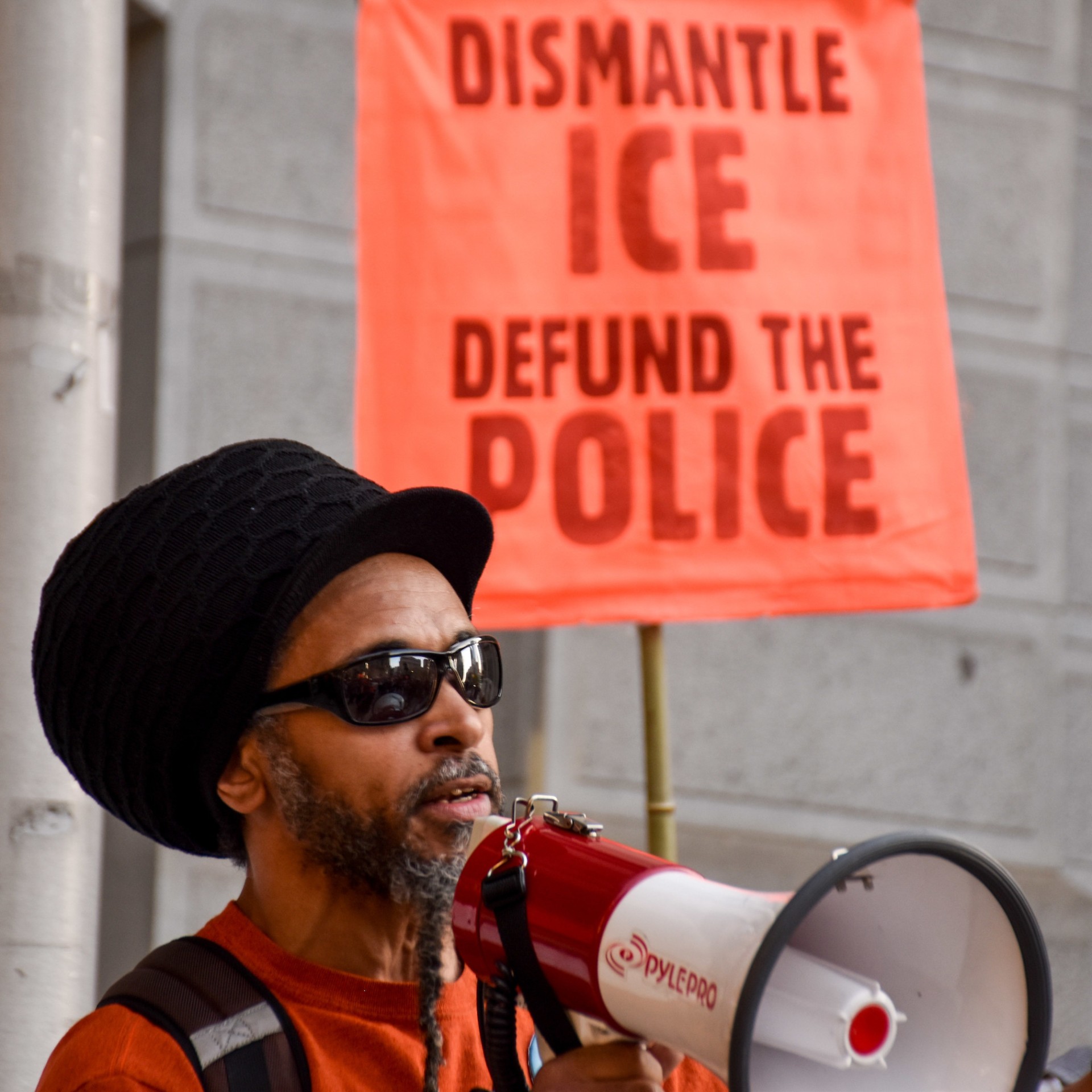 Kempis Songster speaks into a megaphone with a banner behind him that reads 'Dismantle ICE, Defund The Police'