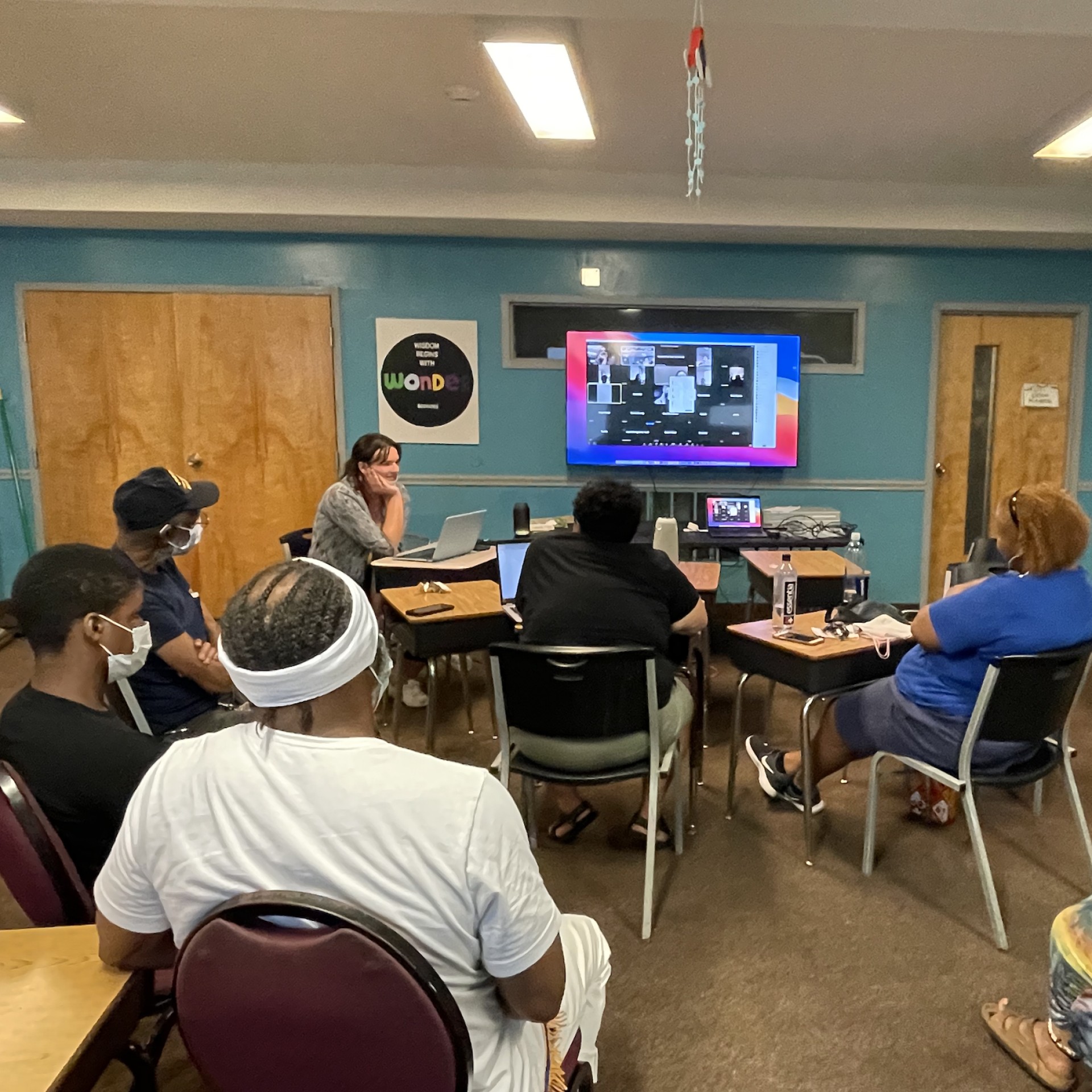 A group of family members of the incarcerated sit in chairs facing a screen where a bunch of people are online to join the in-real life participants in a hybrid meeting