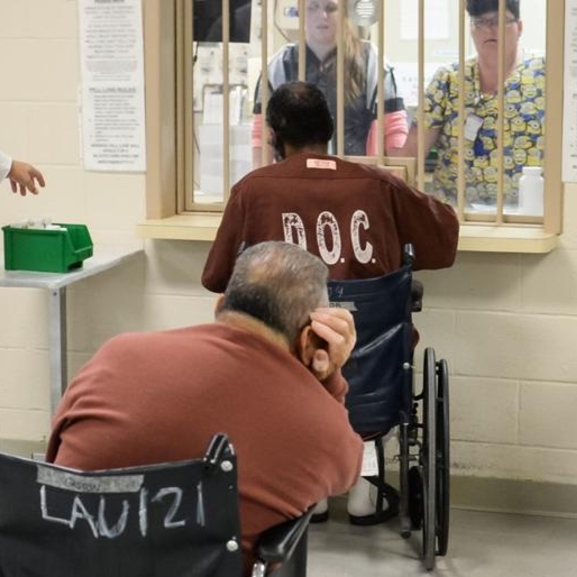 Two incarcerated men in wheel chairs and brown prison clothes wait in a medication line while a man in brown prison pants and a white shirt stands next to them