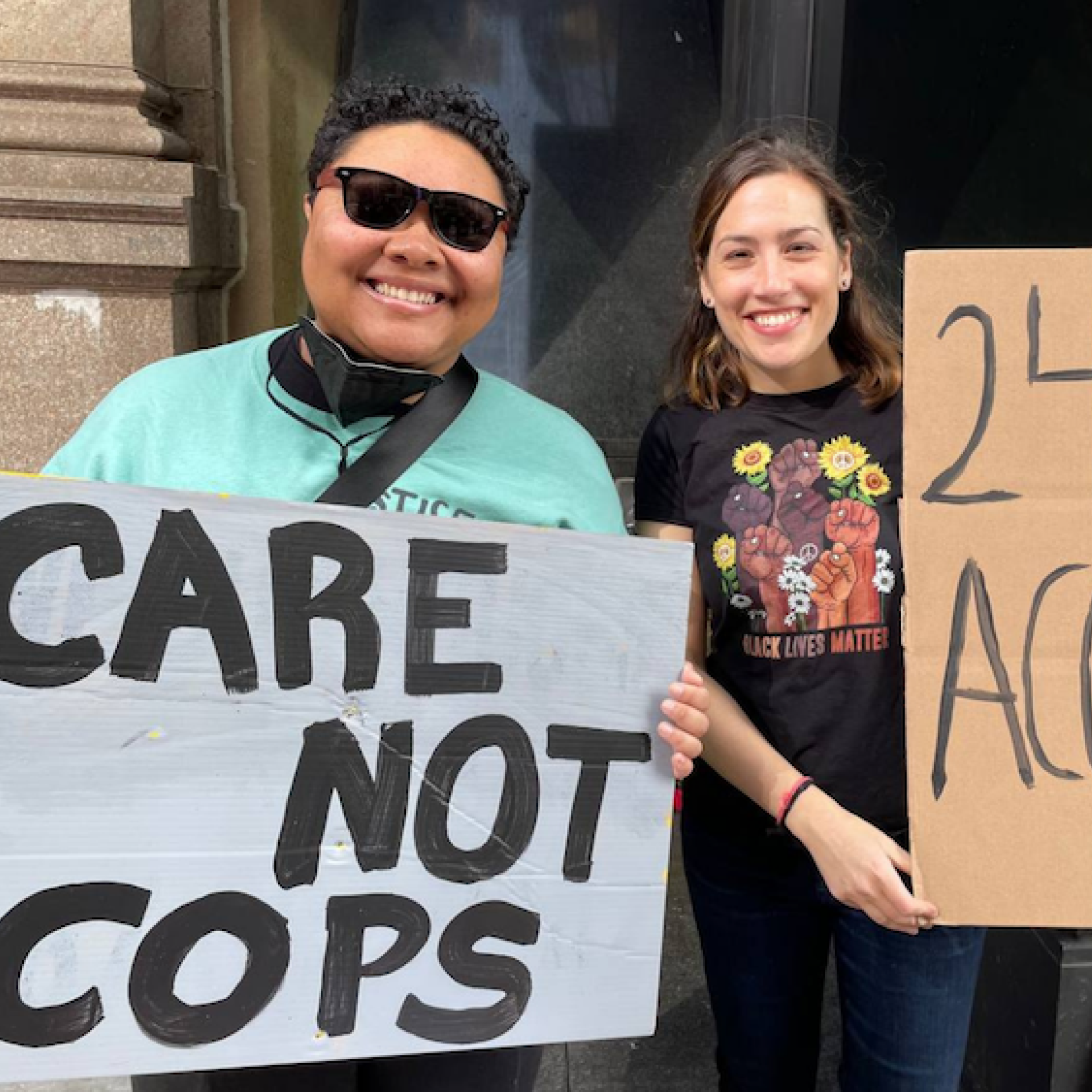 Kris Henderson wears sunglasses and stands next to Nomi Teusch. They are holding signs that say 'Care Not Cops' and '24/7 Access'
