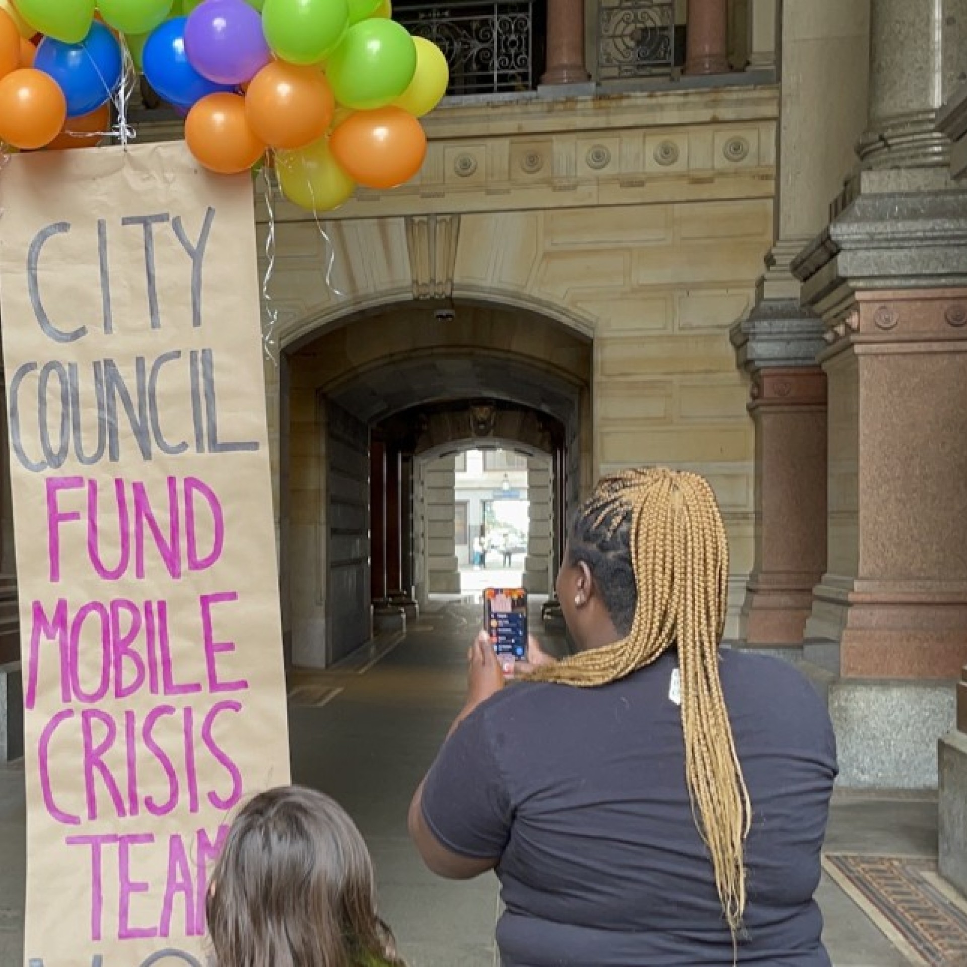 Nikki Grant of Amistad films a colorful balloon banner that reads 'City Council Fund Mobile Crisis Teams'