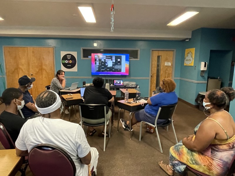 A group of family members of the incarcerated sit in chairs facing a screen where a bunch of people are online to join the in-real life participants in a hybrid meeting