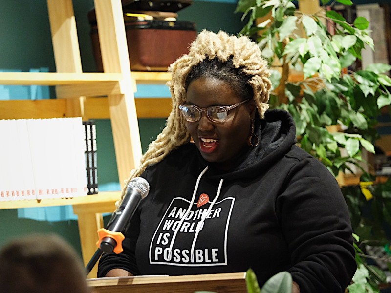 A Black woman with blond hair stands in front of a green plant and speaks into a microphone while wearing a black hooded sweatshirt with the words Another World is Possible on the front of it