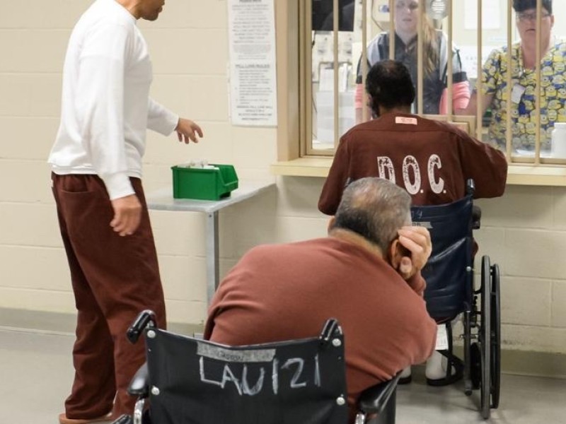 a photo shows two elderly incarcerated people in wheelchairs in line for medication and one incarceraeted person in a white shirt standing next to them