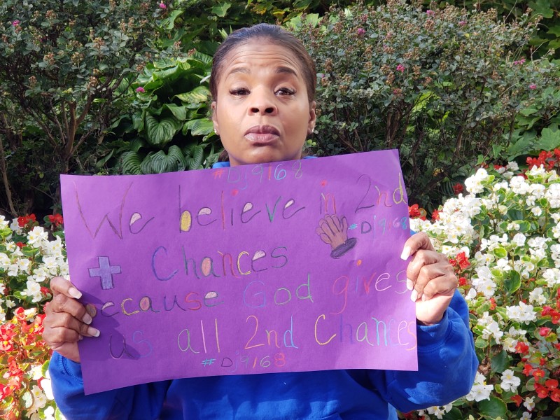 A Black woman looking with determination at the camera holds a purple sigh that says 'I believe in second chancess'