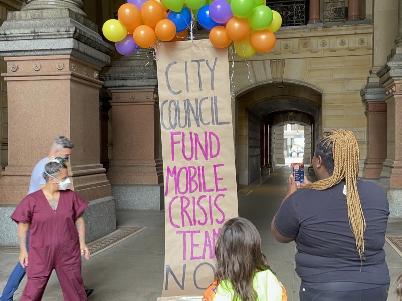 4 people stand around a banner held aloft by colorful balloons. The banner reads "City Council: Fund Mobile Crisis Teams NOW"
