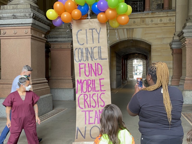 A banner lifted up by balloons reads 'City Council Fund Mobile Crisis Teams Now'