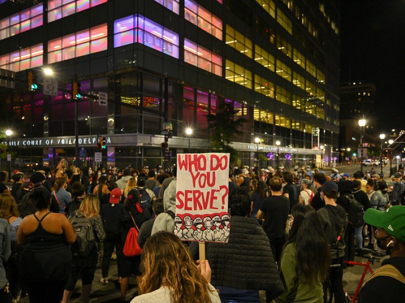 Crowd marches at protest with sign that reads "who do you serve?"