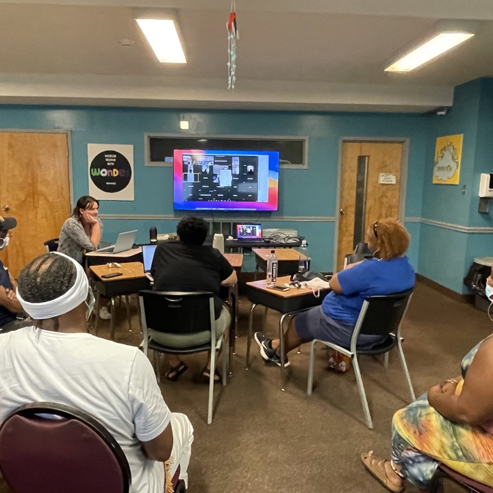 A group of family members of the incarcerated sit in chairs facing a screen where a bunch of people are online to join the in-real life participants in a hybrid meeting
