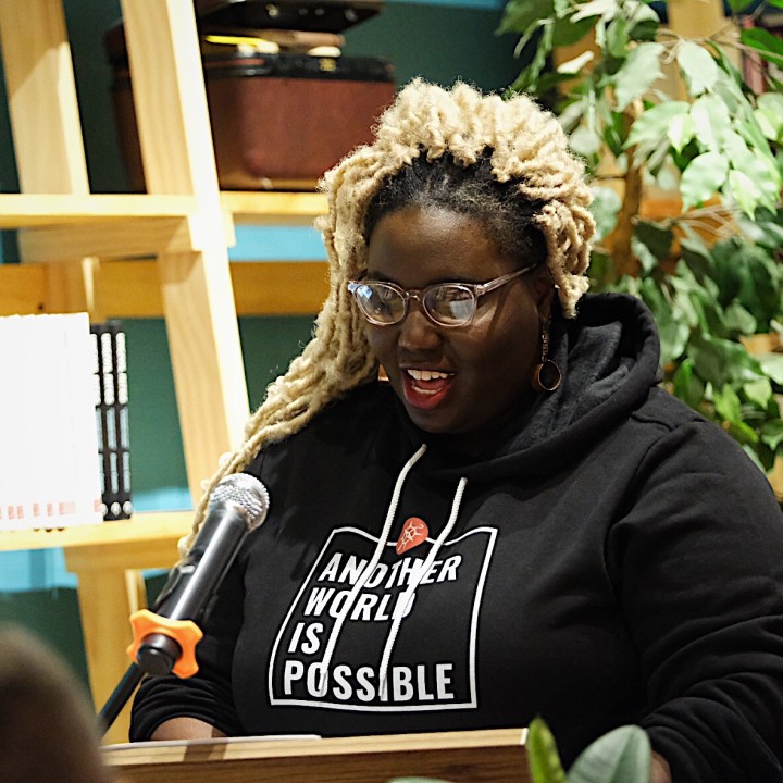 A Black woman with blond hair stands in front of a green plant and speaks into a microphone while wearing a black hooded sweatshirt with the words Another World is Possible on the front of it