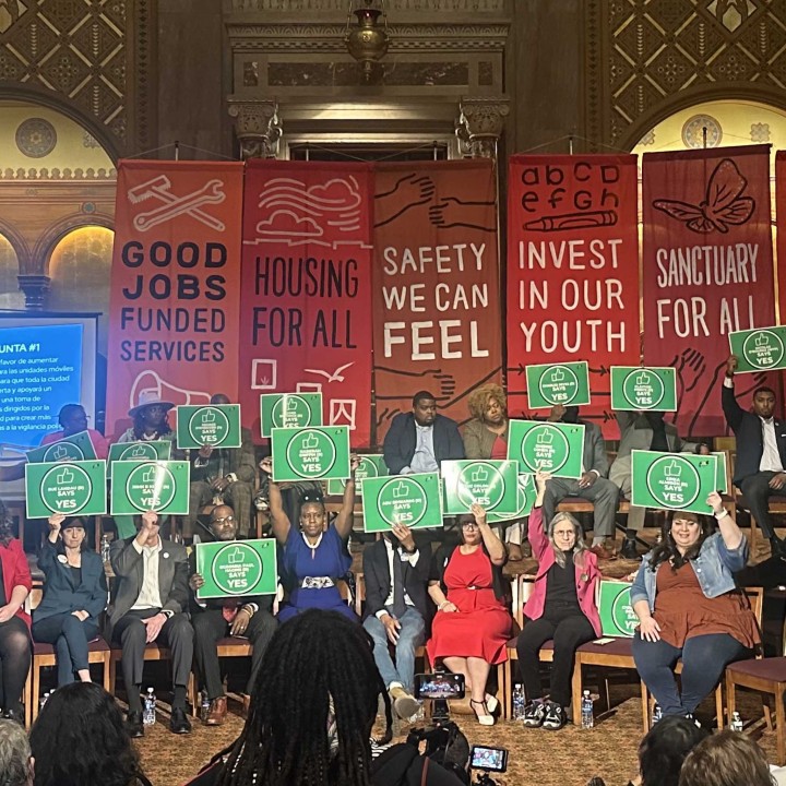 candidates for city council hold up green signs that say yes with red banners behind them 