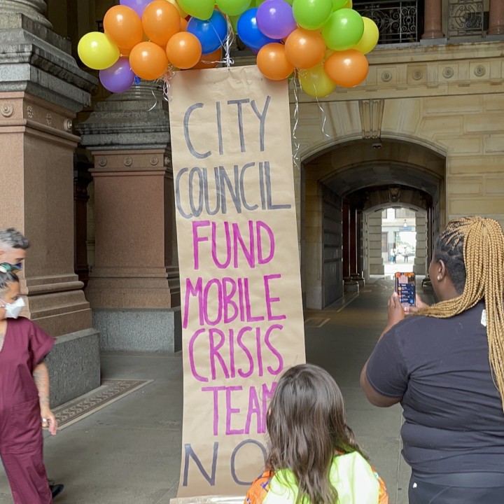 A banner lifted up by balloons reads 'City Council Fund Mobile Crisis Teams Now'
